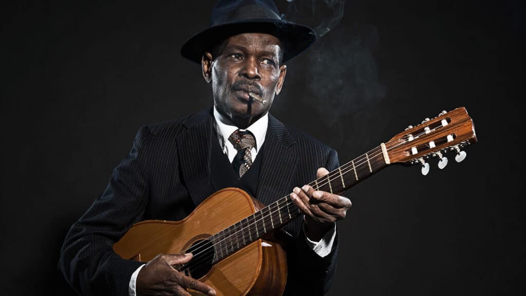 Retro senior afro american blues man. Wearing striped suit with blue hat. Playing acoustic guitar. Smoking cigar.